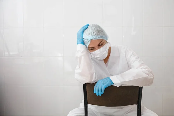 Una joven doctora en traje de protección, gorra, máscara y guantes está sentada cansada en una silla en el consultorio médico. — Foto de Stock
