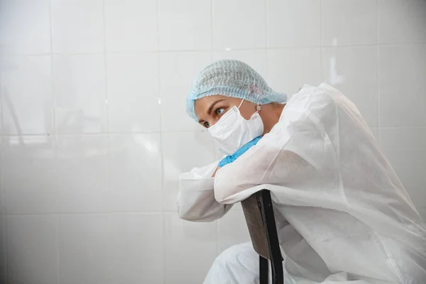 Koruyucu takım elbiseli, şapkalı, maskeli ve eldivenli genç bir bayan doktor, sağlık ofisinde bir sandalyede yorgun bir şekilde oturuyor.