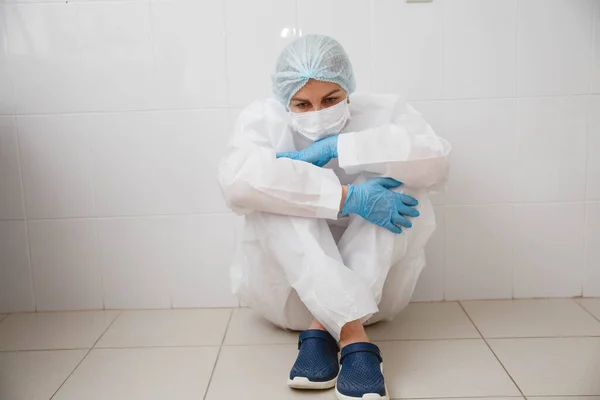 Una joven doctora en traje de protección, gorra, máscara y guantes está sentada cansada en el suelo con las rodillas abrazadas — Foto de Stock