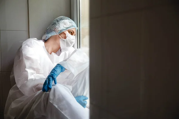 Una joven doctora en traje de protección, gorra, máscara y guantes está sentada cansada cerca de la ventana del alféizar de la ventana — Foto de Stock