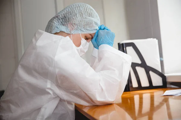 a young female doctor in a protective suit, cap, mask and gloves sits wearily at the table with her eyes closed. High quality photo