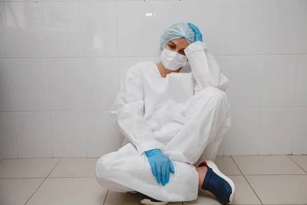 Una joven doctora en traje de protección, gorra, máscara y guantes está sentada cansada en el suelo — Foto de Stock
