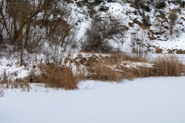 Donmuş taş ocağı yapraksız ağaçlar ve karlı tepelerle çevrili bulutlu bir kış gökyüzü altında, sakin ve geniş bir soğuk mevsim manzarası yaratıyor.