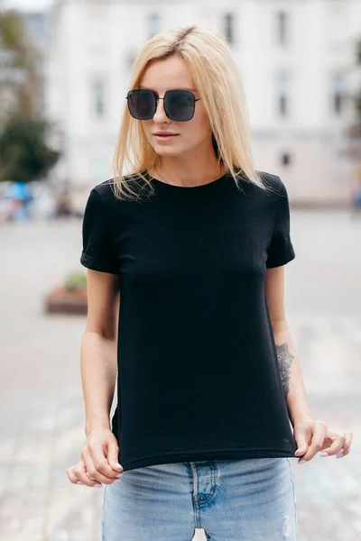 穿着黑色T恤 头戴眼镜 穿着时尚的金发女孩在街上摆姿势 穿着都市风格的服装 街头摄影 — 图库照片