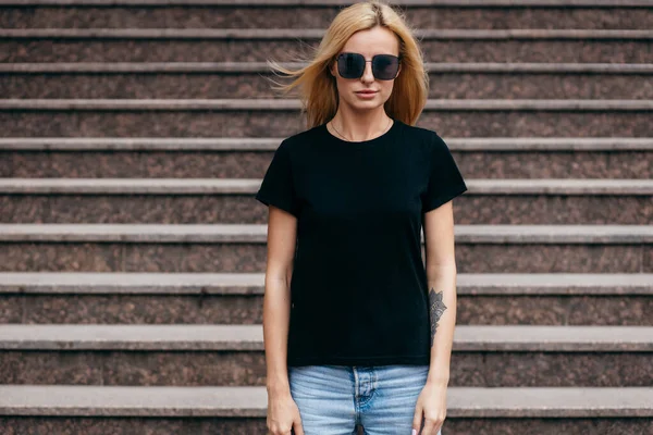 穿着黑色T恤 头戴眼镜 穿着时尚的金发女孩在街上摆姿势 穿着都市风格的服装 街头摄影 — 图库照片