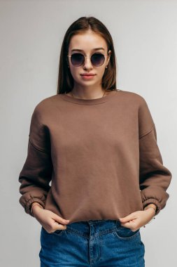 Gözlüklü ve kapüşonlu genç bir kadın stüdyoda beyaz arka planda poz veriyor.