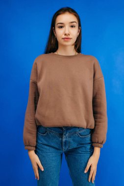 Gözlüklü, tıbbi maskeli ve kapüşonlu genç bir kadın stüdyoda mavi arka planda poz veriyor.