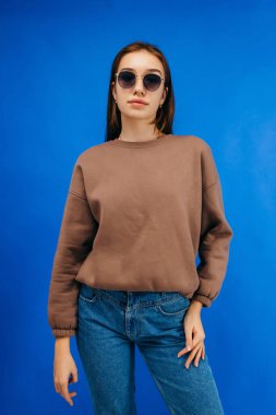 Gözlüklü, tıbbi maskeli ve kapüşonlu genç bir kadın stüdyoda mavi arka planda poz veriyor.