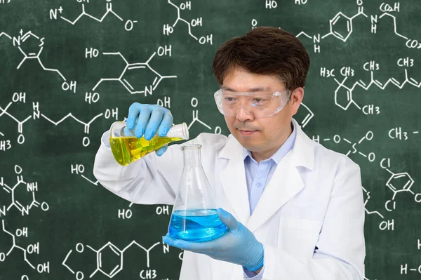 A middle-year Asian male scientist wearing gloves and holding an experimental container in front of a blackboard with a formula written on it.