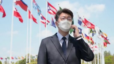 Orta yaşlı Asyalı işadamı maske takıyor ve rüzgarda dalgalanan çeşitli ulusal bayraklar altında akıllı telefon kullanıyor..