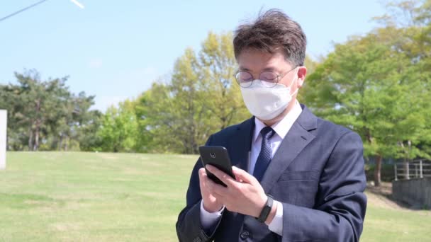 マスクを着用し 芝生の上でスマートフォンを使用している中高年アジアのビジネスマン — ストック動画