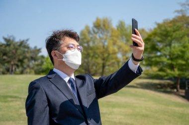 Orta yaşlı Asyalı işadamı maske takıyor ve çimlerde akıllı telefon kullanıyor..