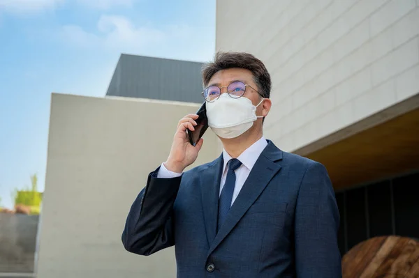 Middle-aged Asian businessman in the city wearing a mask and using a smartphone.