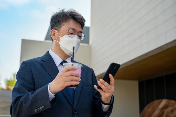 Midaldrende Asiatisk Forretningsmand Byen Iført Maske Holder Kaffekop Bruger Smartphone - Stock-foto