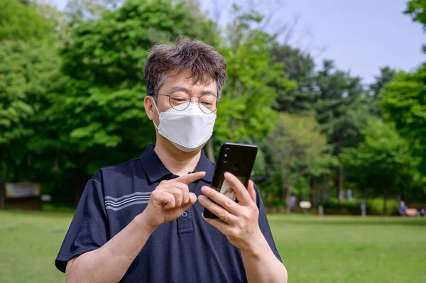 Midaldrende Asiatisk Mand Iført Ansigtsmaske Ved Hjælp Smartphone Plænen - Stock-foto