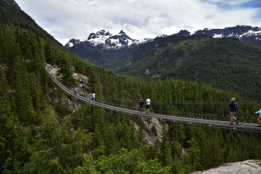 SQUAMISH, BC, CANADA, HAZİRAN 02, 2019: Arkaplanda kar tepeli Gökyüzü Pilot Dağı manzarası olan Askı Köprüsü 'nü geçen insanlar