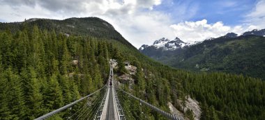 SQUAMISH, BC, CANADA, HAZİRAN 02, 2019: Arkaplanda kar tepeli Gökyüzü Pilot Dağı manzarası olan Askı Köprüsü 'nü geçen insanlar
