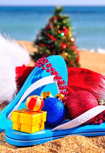 Zapatillas, sombrero de navidad, caja de regalo y árbol de año nuevo en la correa de mar —  Fotos de Stock