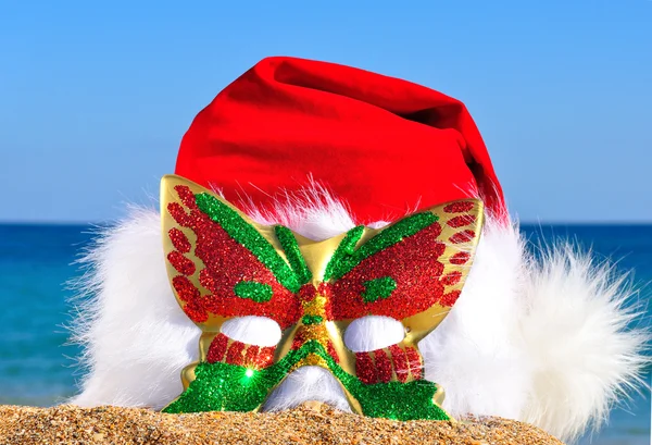 Kerstmuts met glinsterende carnaval masker op de kust — Stockfoto