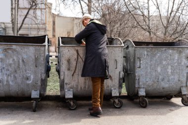 Evsiz genç adam parktaki çöp kutularını karıştırıyor. Evsiz ve işsiz göçmenler sorunu. Sokak serserilerinin yaşam tarzı, sokaklarda yaşamak.