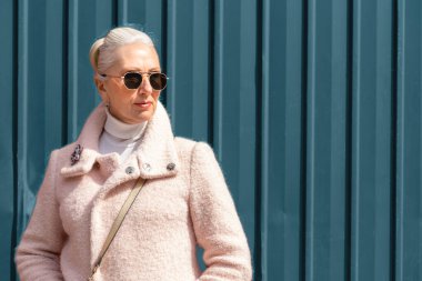 A gray-haired lady in a pink jacket and in black sunglasses stands against a blue metal fence, Woman looks away. Beauty, urban style and fashion concept