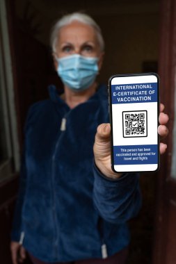 Maskeli orta yaşlı bir kadın elinde uluslararası elektronik koronavirüs aşısı sertifikası ya da gezginler için aşı pasaportu olan bir akıllı telefon tutuyor. Covid-19 'dan dokunulmazlık. Kısıtsız seyahat kavramı