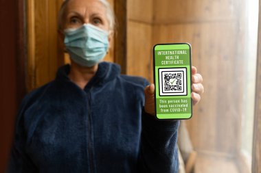 Maskeli orta yaşlı bir kadın elinde uluslararası elektronik koronavirüs aşısı sertifikası ya da gezginler için aşı pasaportu olan bir akıllı telefon tutuyor. Covid-19 'dan dokunulmazlık. Kısıtsız seyahat kavramı