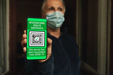 Maskeli kadın cep telefonunda sağlık sertifikası gösteriyor. Bu da kovid 19 'a karşı aşı olduğunu gösteriyor. Seçici bir odak noktası. Kadın telefonda koronavirüs aşısı olduğunu onaylamak için uluslararası bir sağlık belgesi gösteriyor..