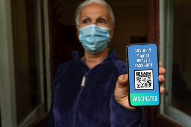 Yüz maskeli yaşlı kadın covid-19 dijital sağlık pasaportu, seçici odaklanma, bulanık bir geçmiş gösteriyor. Koronavirüs salgını sırasında serbest ve güvenli seyahat kavramı