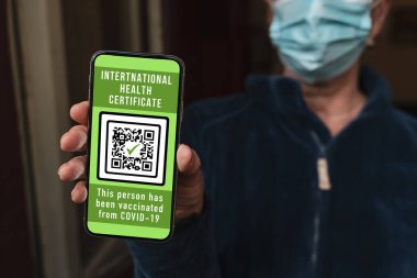 Maskeli kadın cep telefonunda aşı sertifikası gösteriyor. Bu da kovid 19 'a karşı bir aşı olduğunu gösteriyor. Kadın telefonda koronavirüs aşısı olduğunu onaylamak için uluslararası bir sağlık belgesi gösteriyor..