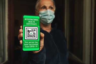 Maskeli kadın cep telefonunda sağlık sertifikası gösteriyor. Bu da kovid 19 'a karşı aşı olduğunu gösteriyor. Seçici bir odak noktası. Kadın telefonda koronavirüs aşısı olduğunu onaylamak için uluslararası bir sağlık belgesi gösteriyor..