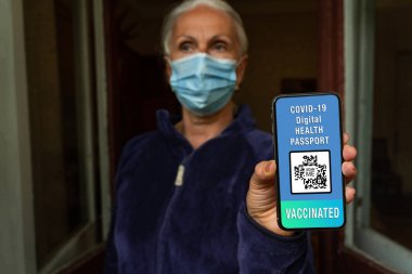 Yüz maskeli yaşlı kadın covid-19 dijital sağlık pasaportu, seçici odaklanma, bulanık bir geçmiş gösteriyor. Koronavirüs salgını sırasında serbest ve güvenli seyahat kavramı