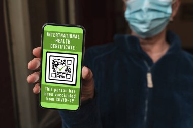Maskeli kadın cep telefonunda aşı sertifikası gösteriyor. Bu da kovid 19 'a karşı bir aşı olduğunu gösteriyor. Kadın telefonda koronavirüs aşısı olduğunu onaylamak için uluslararası bir sağlık belgesi gösteriyor..