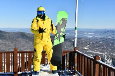 Snowboardcu, güneşli bir günde fotoğraf çekmek için VT 2020 'deki Stowe dağ kayak merkezinin zirvesinde poz veren sarı takım elbiseli. Yüksek çözünürlüklü resim