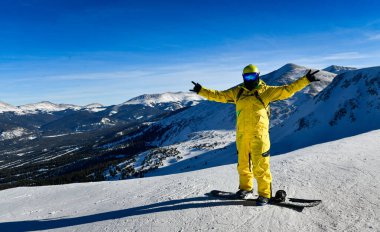 Parlak sarı takım elbiseli snowboardcu, Breckenridge 'in arka planında manzaralı bir manzarayla dağın tepesinde poz veriyor..