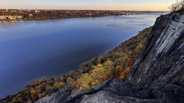 Derin mavi suyun üzerindeki kayalık bir uçurumdan görülen rengarenk sonbahar ağaçlarının panoramik görüntüsü. Sarı, turuncu ve kırmızı tonlarda canlı sonbahar yaprakları çarpıcı mevsimsel bir manzara yaratır..