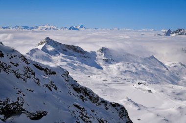 Sersemletici panoramik kar kaplı dağların bulutların üzerinde yükseldiği açık bir kış günü. Engebeli zirveler, yumuşak kar dokuları ve derin mavi gökyüzü dramatik ve dingin yüksek irtifa manzarası yaratır. Seyahat temaları için mükemmeldir.