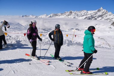 Bir grup kayakçı arka planda çarpıcı alp zirveleriyle karlı bir dağ yamacında duruyor. Renkli kış kıyafetleri içinde kayakçılar aydınlık ve açık bir günde inişlerine hazırlanıyorlar. Her şeyin özünü yansıtan dinamik ve atmosferik bir sahne.
