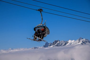 Kayakçılar, arka planda dramatik alp zirveleri olan karlı bir dağ yamacının üzerinde bir teleferiğe biniyorlar. Sahne, kış sporları, yüksek irtifada seyahat ve açık bir günde kayak merkezinin huzurlu ve keyifli atmosferini yakalıyor..