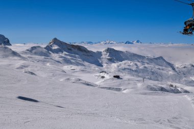 Sersemletici panoramik kar kaplı dağların bulutların üzerinde yükseldiği açık bir kış günü. Engebeli zirveler, yumuşak kar dokuları ve derin mavi gökyüzü dramatik ve dingin yüksek irtifa manzarası yaratır. Seyahat temaları için mükemmeldir.