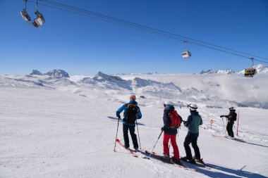 Bir grup kayakçı karlı bir dağ yamacında dikiliyorlar. Sandalye hareket ediyor ve arka planda dramatik alp zirveleri var.. 