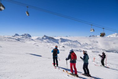 Bir grup kayakçı karlı bir dağ yamacında dikiliyorlar. Sandalye hareket ediyor ve arka planda dramatik alp zirveleri var..