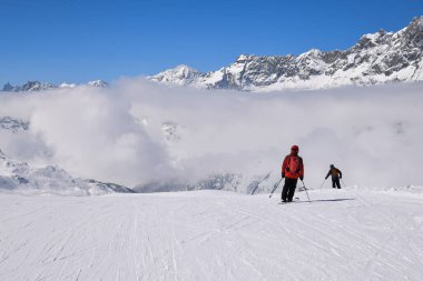 Kayakçılar, bulutların üzerinde yükselen dramatik alp zirveleriyle karlı bir dağ yamacından inerler. Sahne, yüksek rakımlı kış manzaralarının dingin ve görkemli atmosferinin yanı sıra açık hava Alp sporlarının enerjisini de yansıtıyor..
