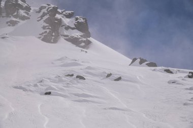 Kış sisinden yükselen karla kaplı kayalık dağ sırtı. Engebeli uçurumlar, yumuşak kar ve düşük görüş mesafesi ile dramatik alp manzarası karamsar, atmosferik bir manzara yaratıyor..