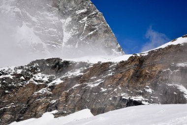 Derin mavi gökyüzünün altında engebeli kaya oluşumlarıyla karla kaplı dağ sırtı. Yokuştan yükselen sisin yüksek dağların gücünü ve güzelliğini yakaladığı dramatik kış manzarası..