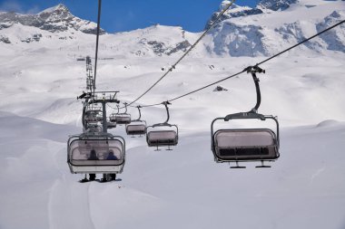 Kayak teleferiği arka planda görkemli dağ zirveleri olan karlı bir dağlık yamacın üzerinde hareket ediyor. Açık havada kış sporu sahnesi, yüksek rakımlı bir kayak merkezinin güzelliğini yakalıyor..