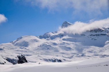 Derin mavi gökyüzünün altında engebeli kaya oluşumlarıyla karla kaplı dağ sırtı. Yokuştan yükselen sisin yüksek dağların gücünü ve güzelliğini yakaladığı dramatik kış manzarası..
