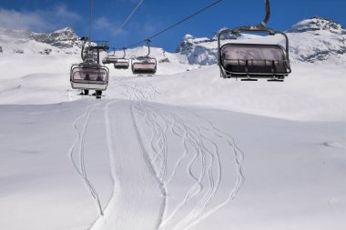 Kayak teleferiği arka planda görkemli dağ zirveleri olan karlı bir dağlık yamacın üzerinde hareket ediyor. Açık havada kış sporu sahnesi, yüksek rakımlı bir kayak merkezinin güzelliğini yakalıyor..