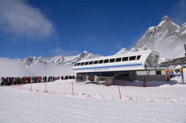 Karlı dağ zirveleri ve açık mavi gökyüzü ile çevrili, aydınlık bir kış gününde modern bir dağ kaldırma istasyonunda bekleyen uzun bir kayakçılar kuyruğu. Yüksek dağlardaki yoğun kayak merkezi atmosferi.