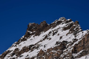 Açık bir kış gününde, derin mavi bir gökyüzünün altında, karla kaplı Alp dağı sırtı. Keskin kayalık tepeler, el değmemiş kar ve gevrek atmosfer kış ve doğa temaları için mükemmel dramatik ve dingin bir yüksek irtifa manzarası yaratır..